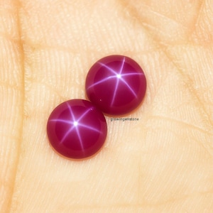 May include: Two polished, deep red gemstones with a six-rayed star effect. The gems are set against a light tan background. The text "glowinggemstone" is visible on the right side of the image. These are likely rubies.