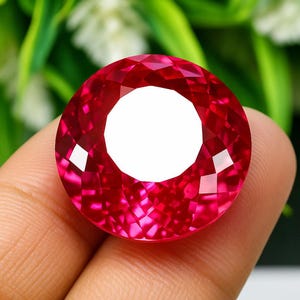 Peut inclure: Une grosse pierre précieuse ronde et facettée, d'un rouge profond et vibrant. La gemme a un centre blanc proéminent et est tenue dans une main. La taille de la pierre crée de nombreuses surfaces réfléchissantes.
