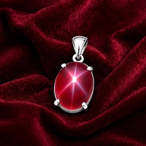May include: A silver pendant with an oval, star-shaped ruby gemstone. The ruby is a deep red color with a six-pointed star effect. The pendant is set against a rich, red velvet background.