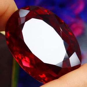 Puede incluir: Una gran gema facetada de forma ovalada con un tono rojo intenso. La gema tiene un corte brillante, que refleja la luz y crea un efecto brillante. La piedra se sostiene entre los dedos, mostrando su tamaño y color.