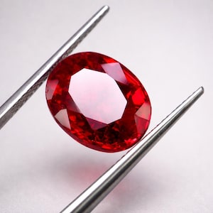 Könnte beinhalten: Ein lebendiger, oval geschliffener Rubin, gehalten von einer silbernen Pinzette vor weißem Hintergrund. Der Rubin zeigt einen tiefroten Farbton mit mehreren Facetten, die das Licht reflektieren. Die Pinzette ist so positioniert, dass die Form und Brillanz des Edelsteins zur Geltung kommen.