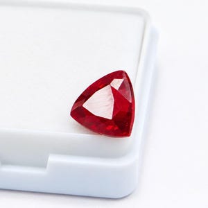 May include: A vibrant red gemstone cut into a triangular shape with multiple facets that reflect light. The stone is positioned on the corner of a white plastic storage container, showcasing its deep crimson colour and sharp, geometric edges.