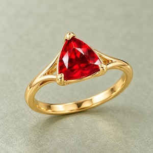Puede incluir: Un anillo de oro con una gema roja de corte triangular engastada en una montura de tres garras. La banda tiene un acabado liso y pulido con un diseño de brazo dividido que se estrecha hacia la piedra central, creando un aspecto equilibrado.