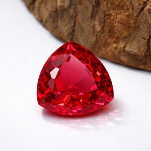 May include: A faceted, triangular-shaped, vibrant red gemstone. The gem has many facets that reflect light, creating a brilliant sparkle. The gemstone is set against a white background with a piece of brown wood in the background.