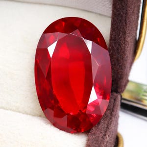 May include: A large, faceted, oval-cut ruby gemstone. The gem is a deep, vibrant red color and is set in a brown velvet jewelry box. The facets catch the light, creating a brilliant display. The ruby is a focal point.
