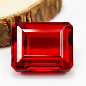 Può includere: Una gemma sfaccettata, rettangolare, rosso intenso con una lucentezza brillante. La gemma ha un design a taglio a gradini, con una serie di sfaccettature parallele. La gemma è posta su uno sfondo bianco e un pezzo di legno.