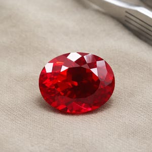 Könnte beinhalten: Ein facettierter, oval geformter, leuchtend roter Edelstein. Der Edelstein liegt auf einem neutralfarbenen Stoff, wobei sich das Licht in seinen vielen Facetten spiegelt. Der Edelstein hat eine tiefe, satte rote Farbe und scheint ein Rubin zu sein.