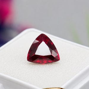 May include: A triangular, deep red gemstone with multiple facets. The gem is set against a white background. The gem's shape is a perfect equilateral triangle, with a clear, open center. The gem has a bright, reflective surface.