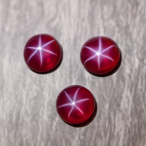 Peut inclure: Trois pierres précieuses rondes et polies, de couleur rouge profond. Chaque gemme présente un effet d'étoile à six rayons, avec des lignes blanches rayonnant du centre. Les pierres sont posées sur une surface neutre et texturée.