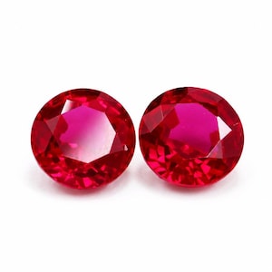 Puede incluir: Dos piedras preciosas redondas, facetadas y de un rojo vibrante con talla brillante. Cada piedra muestra un tono carmesí intenso y múltiples facetas triangulares que reflejan la luz, creando una apariencia brillante y destellante.