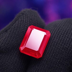 May include: A faceted, rectangular, ruby-red gemstone with a polished surface. The gem is held against a dark gray, textured background, creating a contrast that highlights its vibrant color and geometric shape. The gem's facets catch the light, creating a sparkling effect.