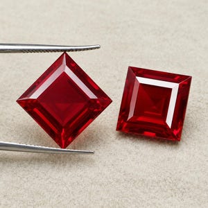 Puede incluir: Dos piedras preciosas rojas vibrantes de corte cuadrado con superficies facetadas. Una piedra es sostenida horizontalmente por pinzas de metal plateado, mientras que la otra se encuentra al lado. Ambas piedras muestran un color carmesí.