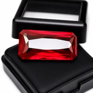 May include: A rectangular, faceted, deep red gemstone with sharp edges and a polished surface, resting on the black velvet-lined interior of an open, black plastic display box. The stone exhibits clear light reflections on its top facets.