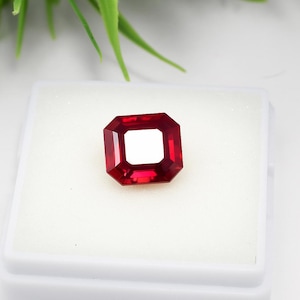 Puede incluir: Una gema de rubí tallada en forma rectangular, de color rojo vibrante. La gema se presenta sobre un fondo blanco, resaltando su claridad y brillo. La piedra preciosa tiene forma octogonal con un centro cuadrado.