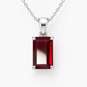 Pode incluir: Um colar de tom prateado com um pendente de pedra preciosa retangular de cor vermelho-escuro, preso por quatro garras. O pendente está suspenso numa delicada corrente prateada ligada a um elo triangular no topo da cravação.