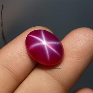 Puede incluir: Gema ovalada de color rojo intenso con un patrón de estrella de seis puntas. El efecto de estrella se crea por la reflexión de la luz en las inclusiones de la piedra. La gema se sostiene en una mano, mostrando su superficie pulida y color vibrante.