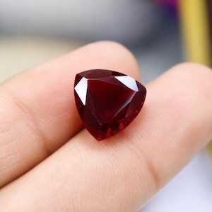 Può includere: Una gemma sfaccettata di colore rosso intenso, tagliata in forma triangolare. La pietra presenta bordi geometrici netti e una superficie lucida che riflette la luce, mostrando una ricca e satura tonalità cremisi in tutta la sua struttura.