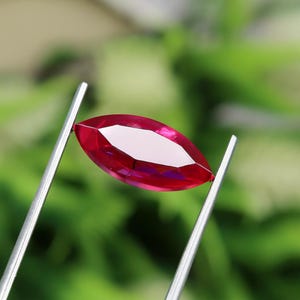 May include: A marquise-cut, faceted ruby gemstone held by silver tweezers. The ruby is a vibrant red color with a bright, reflective surface. The background is a soft, blurred green, suggesting natural foliage.