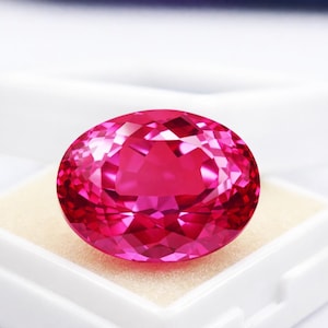 Peut inclure: Une pierre précieuse rose vif de forme ovale et taillée à facettes. La gemme est mise en valeur sur un fond blanc, révélant sa taille brillante et sa couleur intense. La pierre est présentée dans un écrin blanc avec un intérieur beige.