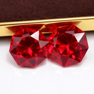 Könnte beinhalten: Zwei facettierte, leuchtend rote Edelsteine mit geometrischem Design. Die Edelsteine werden auf einer weißen Oberfläche mit einem goldenen und braunen Hintergrund präsentiert. Die Edelsteine sind wahrscheinlich für die Schmuckherstellung oder das Sammeln gedacht.