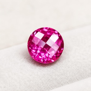 May include: A round, faceted gemstone with a vibrant, saturated hot pink colour. The stone features a precise, symmetrical cut with multiple triangular and quadrilateral facets that reflect light, creating a brilliant, sparkling appearance.
