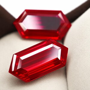 May include: Two vibrant red, faceted gemstones with a hexagonal cut. Each stone features sharp, geometric edges and a polished surface that reflects light, creating bright white highlights across the deep crimson, translucent crystalline structure.