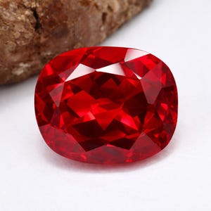 Peut inclure: Une pierre précieuse rouge vif, de forme ovale et à multiples facettes. La gemme présente une grande clarté et reflète la lumière. La pierre est posée sur un fond blanc, avec une roche brune en arrière-plan.