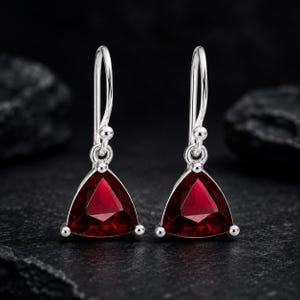Puede incluir: Un par de pendientes colgantes de plata con piedras preciosas de color rojo intenso talladas en forma triangular y facetadas. Cada pendiente cuelga de un gancho de plata curvado, con un pequeño detalle de cuenta de plata en la unión.