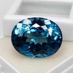 May include: A large, oval-cut gemstone with a deep, vibrant blue colour and a faceted surface that reflects light. The stone features a brilliant, multi-faceted design that creates a sparkling effect across its polished, translucent blue surface.