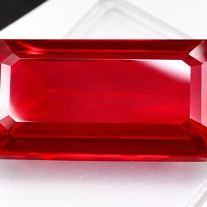 May include: A rectangular, faceted, deep red gemstone. The gem has a clear, polished appearance with sharp edges and a symmetrical shape. The color is a vibrant, saturated red, suggesting a ruby or similar precious stone.