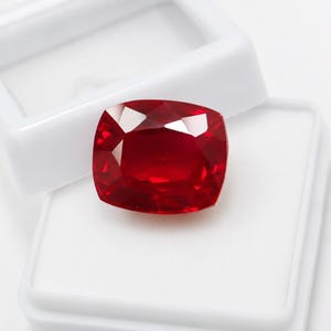 Pode incluir: Uma gema de cor vermelha profunda e vibrante com um corte almofada e múltiplas facetas que refletem a luz. A gema está posicionada centralmente, repousando sobre a base de uma caixa de exibição de plástico branco aberta.