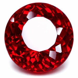 Puede incluir: Una gema roja brillante, probablemente un rubí, de forma redonda y facetada, sobre un fondo blanco. La superficie de la gema refleja la luz, mostrando sus múltiples facetas y su intenso color. La gema es de un rojo profundo y saturado.