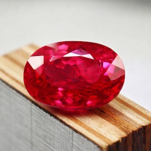 May include: A vibrant, oval-cut ruby gemstone. The gem is a deep, saturated red with multiple facets that reflect light. It rests on a wooden surface, showcasing its brilliance and color. The gemstone is a focal point, highlighting its beauty.