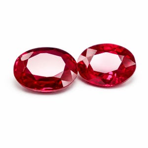 Pode incluir: Duas pedras preciosas de corte oval com uma cor vermelha profunda e vibrante. Cada pedra apresenta uma superfície facetada que reflete a luz, criando uma aparência brilhante e cintilante. As gemas estão posicionadas lado a lado sobre um fundo branco.