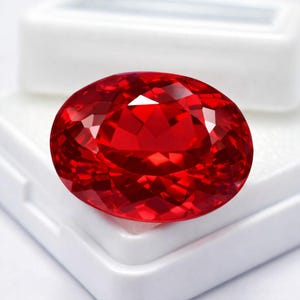 Könnte beinhalten: Ein facettierter, ovalförmiger, leuchtend roter Edelstein. Der Edelstein ist vor einem weißen Hintergrund platziert, der seinen Brillantschliff und seine intensive Farbe hervorhebt. Die Facetten des Edelsteins reflektieren das Licht.