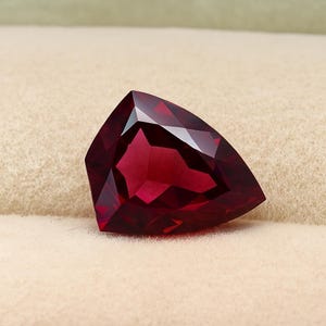 Könnte beinhalten: Ein facettierter, dreieckiger Edelstein in tiefroter Farbe. Der Stein hat viele Facetten, die das Licht reflektieren und einen brillanten Glanz erzeugen. Der Edelstein liegt vor einem neutralen Hintergrund.