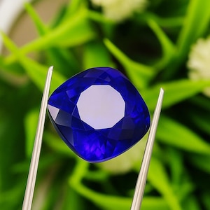 Op de afbeelding: Een diepblauwe, kussenvormig geslepen edelsteen wordt vastgehouden door een zilveren pincet. De facetten van de edelsteen reflecteren het licht, waardoor een briljante weergave ontstaat. De achtergrond is een zachte focus van groen gebladerte.