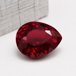 Könnte beinhalten: Ein facettierter, birnenförmiger Rubin-Edelstein. Der Edelstein hat eine tiefe, satte rote Farbe mit mehreren Facetten, die das Licht reflektieren. Der Edelstein ist vor einem weißen Hintergrund platziert, mit einer strukturierten grauen Box im Hintergrund.