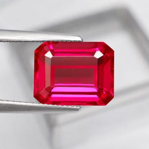 Peut inclure: Une pierre précieuse rectangulaire de taille émeraude d'un rouge profond et saturé. La pierre présente des bords facettés précis qui reflètent la lumière, maintenue par les pointes métalliques argentées d'une pince de bijoutier professionnelle.