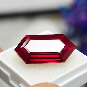 Pode incluir: Uma pedra preciosa de cor vermelho-profundo com facetas e formato hexagonal alongado. A pedra apresenta bordas geométricas nítidas e uma superfície polida que reflete a luz, criando reflexos brancos brilhantes nas facetas superiores.