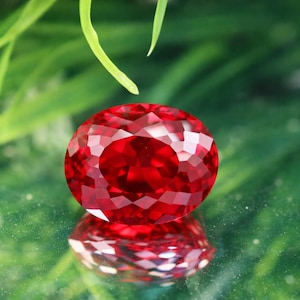 以下が含まれることがあります： 多面的な、楕円形にカットされた、深い赤色の宝石。 宝石は非常に反射性が高く、光を捉える複数のファセットがあります。 背景はぼやけた緑色で、緑の葉が少し見えます。