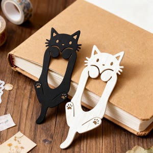 Peut inclure: Deux marque-pages en forme de chat, un noir et un blanc, posés sur un livre fermé. Les marque-pages en bois présentent un design découpé avec des empreintes de pattes et un visage souriant. Accessoire charmant pour les lecteurs.