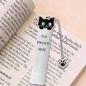 May include: A silver bookmark with a black cat design at the top, a chain, and a heart-shaped charm with a paw print. The bookmark reads "YOU PAUSED HERE" in black text. The bookmark is placed in an open book.