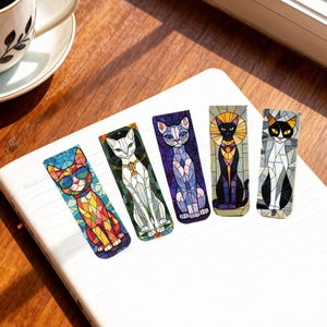 May include: Five colorful cat-themed bookmarks with a stained-glass effect. Each bookmark features a different cat design, including one wearing sunglasses. The bookmarks are arranged on a white notebook, with a coffee cup in the background.