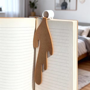 Puede incluir: Un marcapáginas marrón en forma de café derramado, con una pequeña taza de cerámica blanca adjunta. El marcapáginas está insertado en un cuaderno con páginas rayadas. Un accesorio original para los lectores.