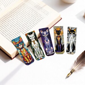 May include: Five colorful cat-themed bookmarks with stained glass designs are displayed next to an open book. The bookmarks feature various cat poses and colors, including orange, white, purple, black, and gray. A feather quill rests nearby.