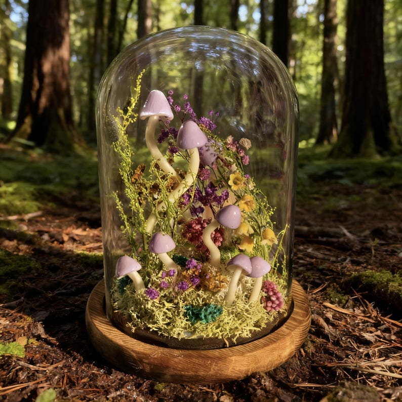 Pu&ograve; includere: Un terrario a campana di vetro con sculture di funghi lavanda in miniatura tra fiori secchi e muschio. Il terrario poggia su una base di legno rotonda, creando una scena boschiva stravagante. Lo sfondo &egrave; una foresta sfocata.