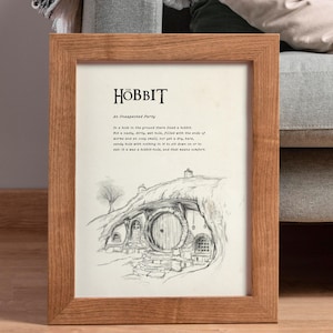 Op de afbeelding: Een ingelijste print met een potloodtekening van een hobbitgat. De print bevat de titel "The Hobbit" en een citaat uit het boek. De lijst is gemaakt van lichtbruin hout en de print wordt op een houten vloer gepresenteerd.