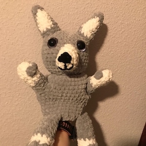 May include: A handmade gray and white crochet kangaroo plush toy with large black eyes and a black nose. The kangaroo has its arms and legs outstretched, with white accents on its ears, paws, and feet. The toy is soft and cuddly.