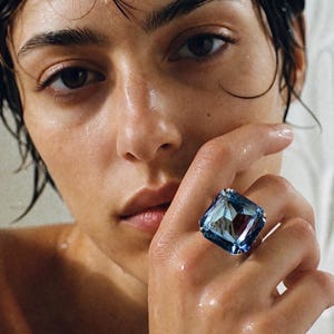 May include: A large, square-cut, light blue gemstone ring featuring a faceted surface that reflects light. The ring is set on a silver-toned band and is worn on a finger, showcasing the prominent size and clear, icy blue colour of the stone.